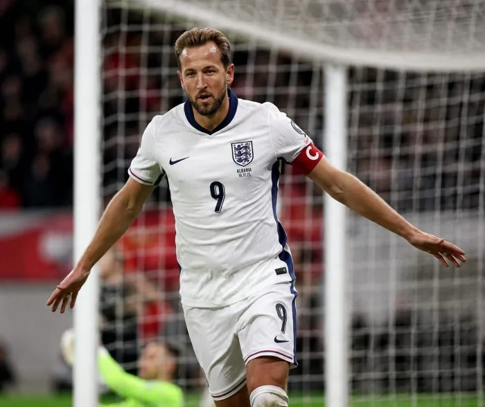 A high-resolution horizontal photograph of England captain Harry Kane celebrating a goal during the 2026 FIFA World Cup. He is running across the pitch with open arms, wearing the white England national jersey, with an ecstatic crowd in the stadium background under vibrant stadium floodlights.