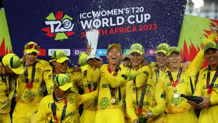 A wide-angle horizontal sports photograph showing members of the Australian women's cricket team celebrating on the historic green outfield at Lord's under bright daylight, with confetti falling and a cheering stadium crowd blurred in the background.