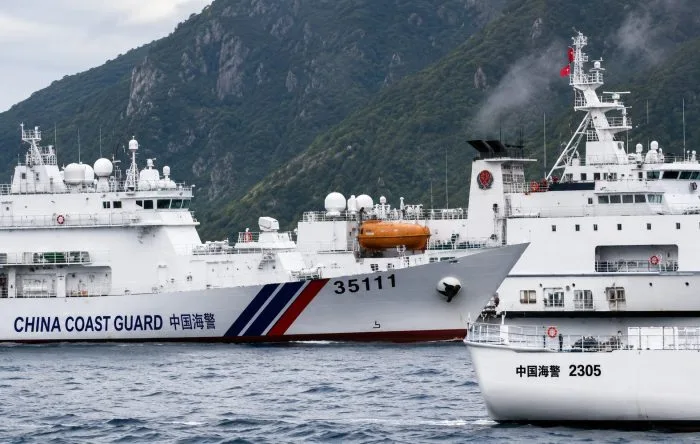A horizontal, wide-angle photograph capturing an official maritime coast guard vessel navigating choppy ocean waters under a vast, overcast sky, with a rocky, uninhabited island visible on the horizon.