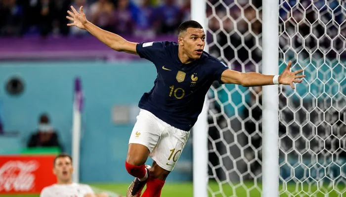 A high-resolution horizontal photograph of Kylian Mbappé celebrating his second goal at MetLife Stadium in New Jersey. He is running toward the corner flag with his trademark arms-crossed pose, smiling brightly, while his teammate Michael Olise runs up to congratulate him in front of a cheering crowd clad in blue.