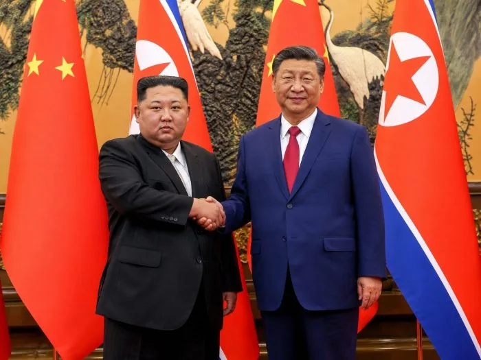 A high-resolution horizontal photograph of Chinese President Xi Jinping and North Korean leader Kim Jong Un standing side-by-side on a red carpet at Kim Il Sung Square in Pyongyang. They are reviewing an official military guard of honor under bright daylight, with large crowds in the background waving national flags of China and North Korea.