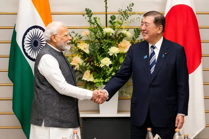 A wide-angle, horizontal photograph of Indian Prime Minister Narendra Modi and Japanese Prime Minister Sanae Takaichi standing side-by-side at a formal press podium during a joint statement, smiling and gesturing warmly, with the national flags of India and Japan displayed prominently behind them.